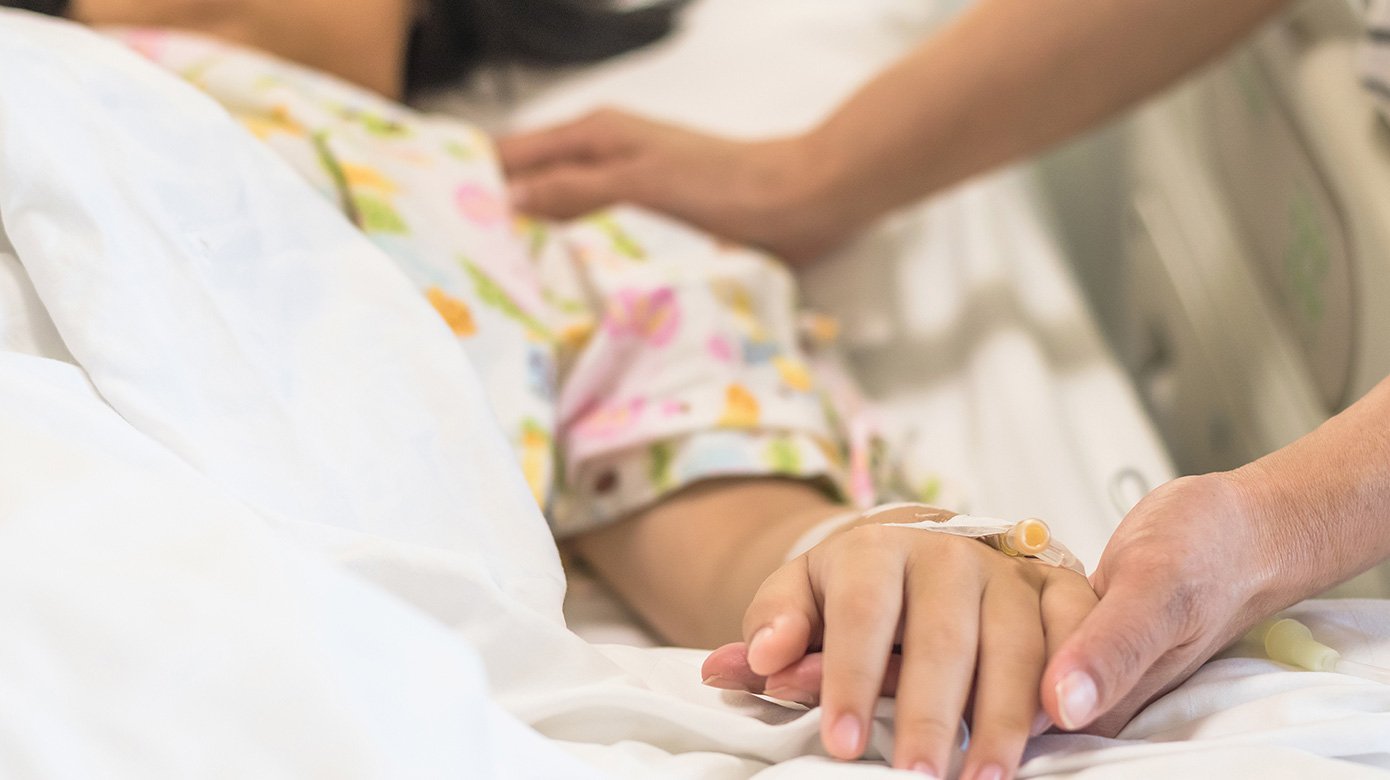 child in hospital bed with caregiver holding her hand